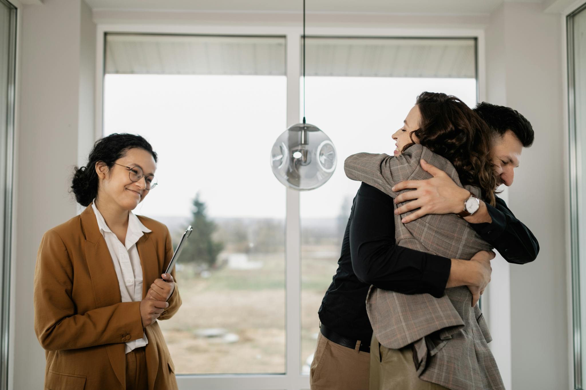 A couple joyfully embraces in their new home as a satisfied real estate agent watches.